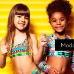 Duas meninas sorrindo vestindo biquínis coloridos com fundo amarelo listrado. Texto: Moda Praia Infantil.