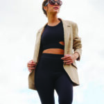 Mulher usando conjunto fitness preto com blazer bege e óculos escuros ao ar livre.