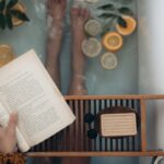 Pés de mulher em banheira com frutas cítricas e livro na mão.